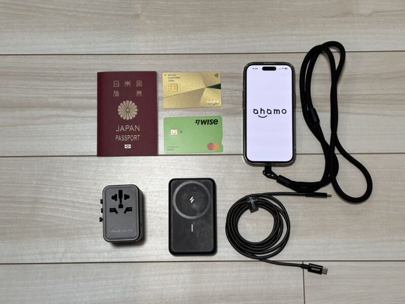 海外旅行の必須の持ち物6つ。パスポート、クレジットカードとWiseカード、ahamoを表示したスマホとネックストラップ、変換プラグ、モバイルバッテリーとUSB-Cケーブル