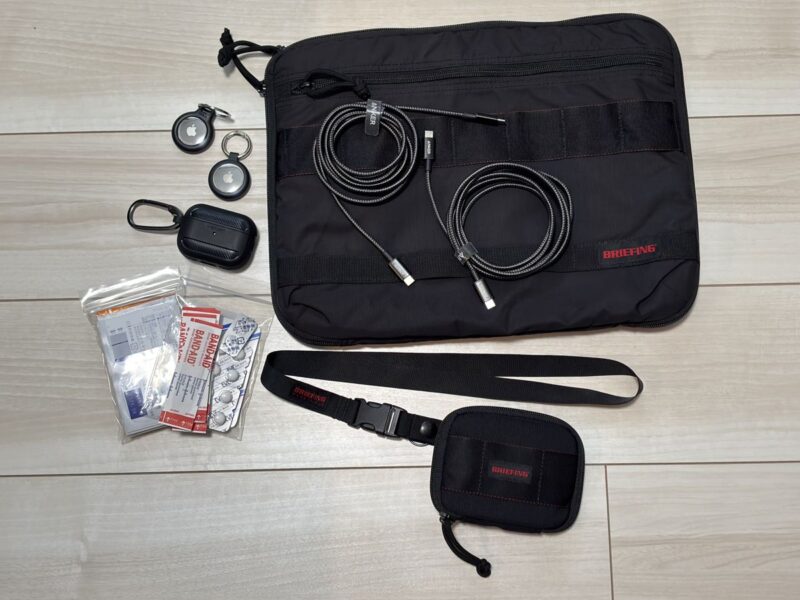 海外旅行であると安心な持ち物。AirTag2個、ケース入りのイヤホン、常用薬と絆創膏、BRIEFINGのPCケースとUSB-Cケーブル2本、NECK HOLDERと小さい財布